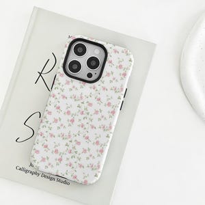 Könnte beinhalten: Eine weiße Handyhülle mit einem floralen Muster aus kleinen rosa Rosen und grünen Blättern. Die Hülle hat einen schwarzen Rand um die Kameralinsen und schwarze Seitentasten. Die Handyhülle liegt auf einem Buch mit dem Text "Calligraphy Design Studio".