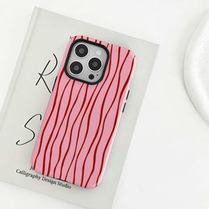 Könnte beinhalten: Rosa Handyhülle mit einem wellenförmigen roten Muster. Die Hülle, mit schwarzem Kamerarahmen, liegt auf einem Buch mit dem Text "Ri S" und "Calligraphy Design Studio".