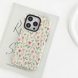May include: A phone case featuring a floral design of pink tulips and green leaves on a cream backdrop. The case has a black camera module and is placed on a book with the text "Calligraphy Design Studio".