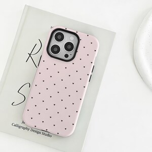 May include: A pink phone case with a black camera module and a pattern of small black hearts. The case is resting on a book with the text "Ri S" and "Calligraphy Design Studio".