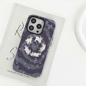 Pode incluir: Uma capa de telefone azul escuro com um padrão de coelhos brancos em círculo, rodeados por estrelas brancas e linhas onduladas. A capa tem um módulo de câmera preto e o texto "Calligraphy Design Studio".