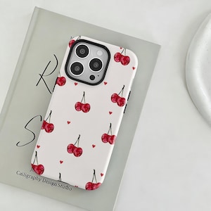 Könnte beinhalten: Weißes Handycase mit einem Muster aus roten Kirschen und kleinen roten Herzen. Das Case hat einen schwarzen Kamerarand und den Text "Calligraphy Design Studio" unten.