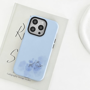 Puede incluir: Una funda de teléfono azul claro con un borde negro para la cámara. La funda presenta un diseño de acuarela de una tortuga y una ballena. La funda del teléfono está sobre un libro con el texto "Calligraphy Design Studio".