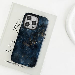 May include: A smartphone case with a dark blue and gold galaxy design. The case has a black border and a camera cutout. The case is resting on a white book with the text "R S" and "Calligraphy Design Studio".