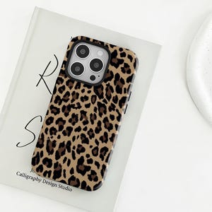 May include: A smartphone case featuring a tan, brown, and black leopard print pattern. The case has a rectangular cutout for a triple-lens camera system on the back. The design covers the entire rear surface of the phone case with a repeating animal spot motif.