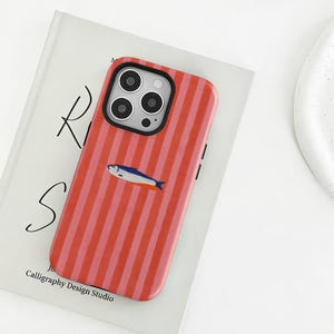 Könnte beinhalten: Eine rot-rosa gestreifte Handyhülle mit einer Illustration eines blau-orangen Fisches. Die Hülle liegt auf einem Buch mit den Buchstaben "R" und "S". Das Buch trägt den Text "Calligraphy Design Studio".