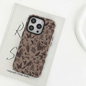 May include: A brown phone case with a black camera module. The case has a repeating pattern of bats, butterflies, mushrooms, witch hats, and other spooky elements. The text "Calligraphy Design Studio" is visible.