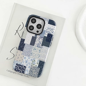 May include: A phone case with a patchwork design in shades of blue and white, featuring floral patterns. The case has a black camera module and the text "Calligraphy Design Studio".