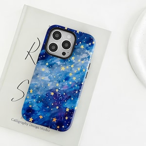 Könnte beinhalten: Eine blaue Handyhülle mit Sternennacht-Design. Die Hülle hat einen dunkelblauen Hintergrund mit Aquarelleffekt und ist mit goldenen Sternen verziert. Die Handyhülle liegt auf einem weißen Buch.