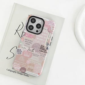 Op de afbeelding: Een telefoonhoesje met een collage-ontwerp met religieuze citaten en bloemaccenten. De hoes heeft een roze en wit kleurenschema met het woord "JESUS" prominent weergegeven. De hoes ligt op een wit boek.