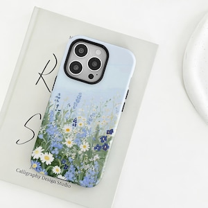 Könnte beinhalten: Hellblaue Handyhülle mit floralem Design mit weißen Gänseblümchen, blauen Wildblumen und grünem Laub. Die Hülle hat einen schwarzen Kamerarahmen und den Text "Calligraphy Design Studio".