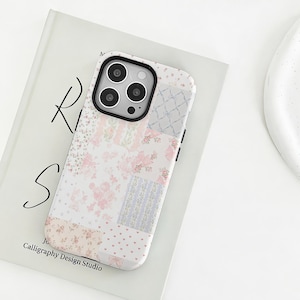 以下が含まれることがあります： ピンク、白、ラベンダーの花柄パッチワークデザインの携帯電話ケース。ケースには黒いカメラフレームが付いています。「Calligraphy Design Studio」の文字が見えます。