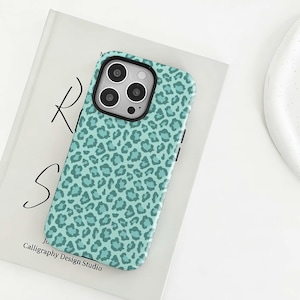 Könnte beinhalten: Eine türkisfarbene Handyhülle mit schwarzem Kamerarand und Leopardenmuster. Die Hülle liegt auf einem Buch mit dem Text "RS" und "Calligraphy Design Studio".