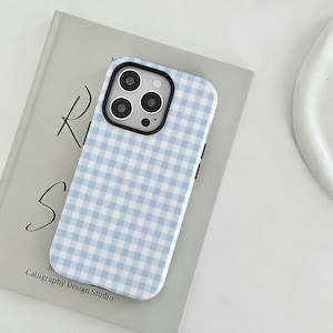 May include: A light blue and white gingham patterned phone case with a black camera border. The phone case is resting on a book with the text "Ri S" and "Calligraphy Design Studio".