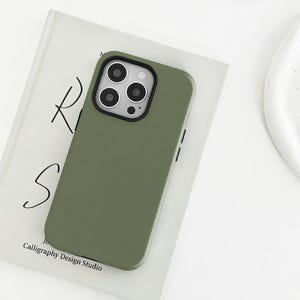 Könnte beinhalten: Olivgrüne Handyhülle mit schwarzem Kamerarahmen. Die Hülle liegt auf einem weißen Buch mit dem Text "RS" und "Calligraphy Design Studio". Die Handyhülle hat eine glatte, matte Oberfläche.