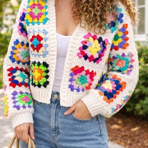 May include: Cream-colored crochet cardigan with colorful square patterns. The cardigan features a V-neck and long sleeves, with vibrant squares in shades of pink, yellow, blue, and green. The garment is worn with blue jeans.