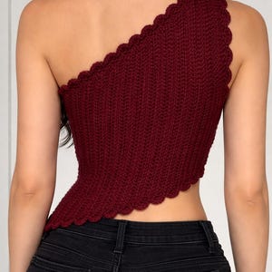 May include: A burgundy one-shoulder top with a scalloped edge. The knitted top is paired with black denim jeans. The top's design features a textured, ribbed pattern.