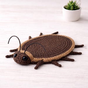 May include: A handmade crochet insect in shades of brown and tan. The insect has large black eyes, curved antennae, and multiple legs. The crochet work is detailed, with a textured appearance. The insect is lying on a light-colored surface.