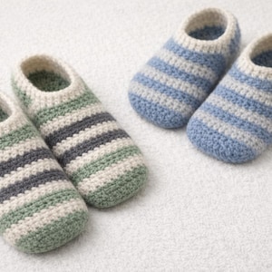 May include: Two pairs of handmade crochet slippers. One pair features green, gray, and cream stripes, while the other has blue and cream stripes. The slippers are designed for indoor wear and are placed on a white surface.