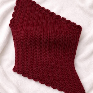 May include: A burgundy one-shoulder crochet top with a scalloped edge. The top has vertical ribbing and an asymmetrical design. The garment is displayed on a white, textured surface, highlighting its rich colour and texture.