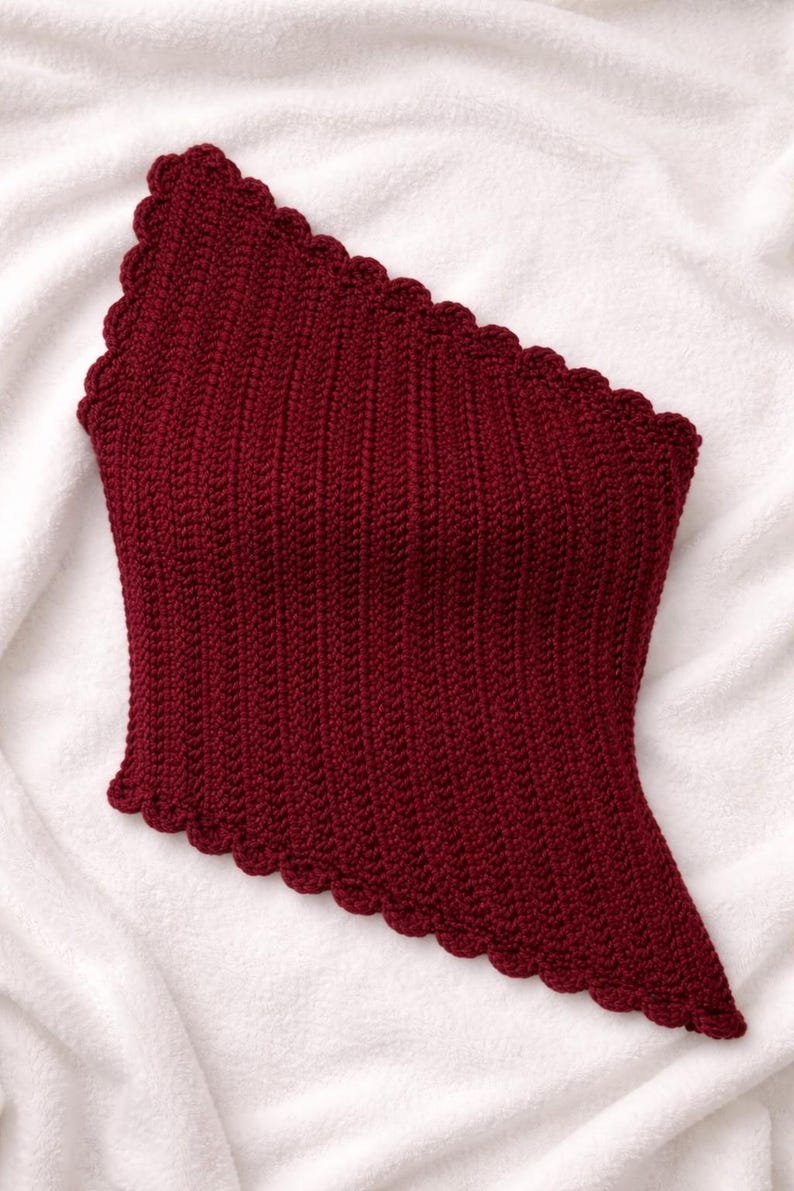 May include: A burgundy one-shoulder crochet top with a scalloped edge. The top has vertical ribbing and an asymmetrical design. The garment is displayed on a white, textured surface, highlighting its rich colour and texture.