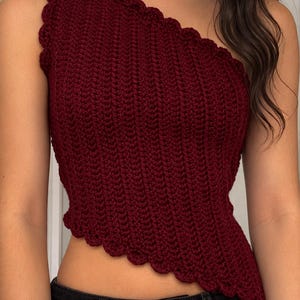 May include: A burgundy crochet one-shoulder top with a scalloped edge. The top is made with a textured stitch and has a fitted silhouette. The garment is paired with black denim jeans.