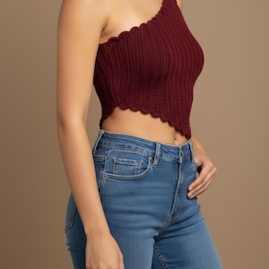 May include: A burgundy one-shoulder crop top with a scalloped edge. The top is knitted and worn with blue jeans. The jeans have a classic five-pocket design.