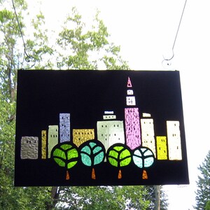 May include: A black framed cityscape art piece with a pink, yellow, and blue building design. The buildings are surrounded by green trees.