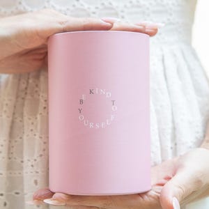 Puede incluir: Un recipiente cil&iacute;ndrico rosa claro sostenido por dos manos. El recipiente tiene las palabras "KIND TO YOURSELF" en blanco, dispuestas en c&iacute;rculo. El fondo es una tela blanca con un dise&ntilde;o de ojal.