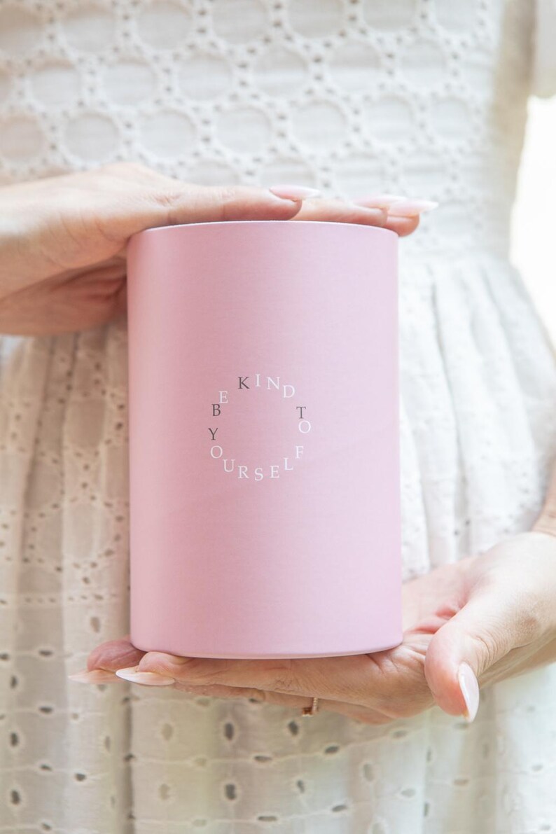 Puede incluir: Un recipiente cil&iacute;ndrico rosa claro sostenido por dos manos. El recipiente tiene las palabras "KIND TO YOURSELF" en blanco, dispuestas en c&iacute;rculo. El fondo es una tela blanca con un dise&ntilde;o de ojal.