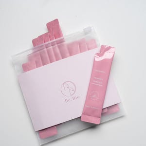 Puede incluir: Un paquete de sobres de suplementos rosas en una bolsa de pl&aacute;stico transparente, con una tarjeta rosa claro y un solo sobre en el exterior. La tarjeta tiene un logotipo y el texto "Re:Bio". El sobre individual tiene texto y un sello.