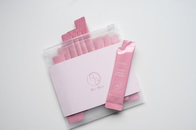 Puede incluir: Un paquete de sobres de suplementos rosas en una bolsa de pl&aacute;stico transparente, con una tarjeta rosa claro y un solo sobre en el exterior. La tarjeta tiene un logotipo y el texto "Re:Bio". El sobre individual tiene texto y un sello.