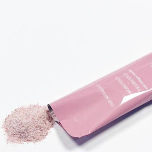 Puede incluir: Un sobre rosa de polvo con el texto "SYNBIOTICS PROBIOTICS with Collagen Peptide" y un peque&ntilde;o mont&oacute;n de polvo rosa que se derrama. El sobre est&aacute; etiquetado "THE MOMENT". El polvo parece ser un suplemento para la salud.