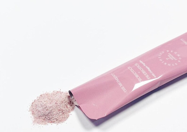 Puede incluir: Un sobre rosa de polvo con el texto "SYNBIOTICS PROBIOTICS with Collagen Peptide" y un peque&ntilde;o mont&oacute;n de polvo rosa que se derrama. El sobre est&aacute; etiquetado "THE MOMENT". El polvo parece ser un suplemento para la salud.