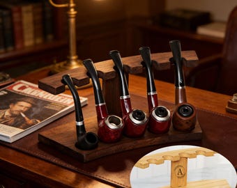 May include: A dark wood pipe stand featuring five slots for tobacco pipes, with a curved base and a central support pillar. An inset circular image shows a light wood version of the same stand with a circular logo containing the letter A.