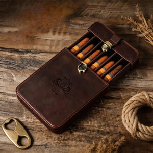 May include: A dark brown leather cigar case with a brass latch closure, containing five cigars with orange and black bands. The case features an embossed logo with the name Anabel. A separate brass cigar cutter is positioned to the left.