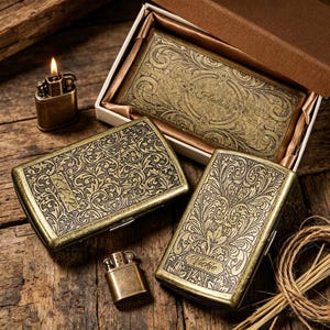 May include: An antique brass-colored lighter with a lit flame sits next to two matching cigarette cases and a rectangular box. The cases and box feature ornate floral patterns and the names "Vickie" and "Haudlets". The items are displayed on a wooden surface.