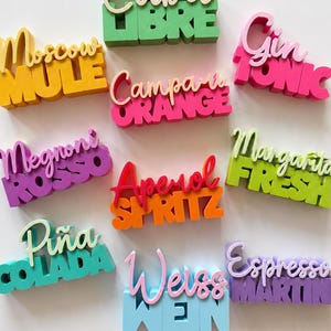 Puede incluir: Una colección de diez letreros impresos en 3D con nombres de cócteles en varios colores y fuentes. Cada letrero muestra el nombre de una bebida como Cuba Libre, Moscow Mule, Gin Tonic, Campari Orange, Negroni Rosso, Aperol Spritz, Margarita Fresh, Piña Colada, Weiss Wein y Espresso Martini.