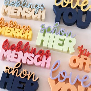 Op de afbeelding: Een verzameling kleurrijke 3D houten woorduitsparingen. De woorden zijn onder meer "lieblings MENSCH", "schon HIER", "love YOU" en "für off". De uitsparingen zijn in verschillende kleuren, waaronder blauw, geel, roze en oranje.