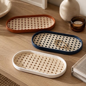 Könnte beinhalten: Drei ovale Tabletts mit erhöhtem Rand in Terrakotta, Marineblau und Weiß. Jedes Tablett verfügt über einen Boden aus geflochtenem Rohrgeflecht mit einem sich wiederholenden achteckigen Muster. Die Tabletts sind auf einer flachen Oberfläche angeordnet.