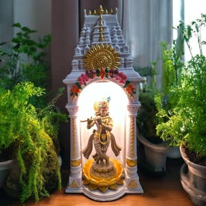 Puede incluir: Una estatua decorativa blanca y dorada de una deidad tocando la flauta, ubicada dentro de una estructura similar a un templo. La estatua está iluminada desde adentro y rodeada de coloridos acentos florales. La estatua está sobre una superficie de madera, con plantas en macetas en el fondo.