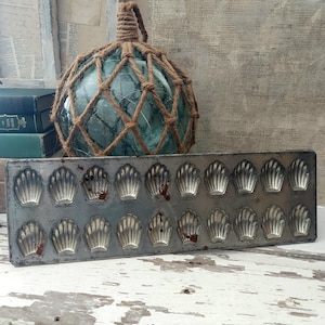 May include: An antique metal madeleine pan with 18 shell-shaped molds sits in front of a vintage glass fishing float encased in a woven rope net. The float is a sea green color.