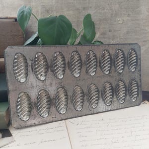 May include: A rectangular metal baking mold featuring sixteen individual oval-shaped cavities with a distinct ribbed, spiral pattern. The mold has a weathered, dark grey finish and is arranged in two horizontal rows of eight indentations each.