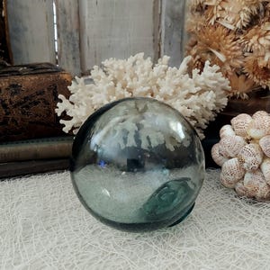 Op de afbeelding: Een heldere, ronde glazen dobber met een groene basis, die de omringende objecten weerspiegelt. De dobber ligt op een wit net, met witte koraal en schelpen op de achtergrond. Decoratief nautisch item.