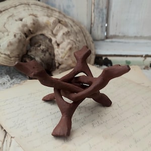 May include: A small, handcrafted, brown wooden stand with a tripod base. The stand is designed to hold a small object. The stand is made of interlocking pieces of wood. The background includes a large, light-colored, organic shape and vintage paper.