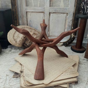 May include: A handcrafted, brown wooden display stand with a unique, open design. The stand features a central vertical support and multiple curved arms extending outwards. It rests on a stack of aged paper, suggesting a vintage aesthetic.