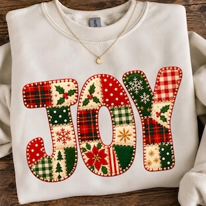 May include: A white crewneck sweatshirt featuring the word JOY printed in large letters. The letters are filled with a patchwork design of red, green, and cream patterns including holly, poinsettias, snowflakes, plaid, and polka dots.