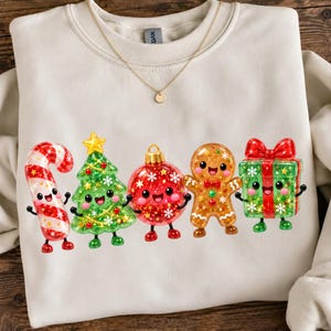 May include: A cream-coloured crewneck sweatshirt featuring a graphic print of five smiling Christmas characters: a red and white candy cane, a green Christmas tree, a red ornament, a gingerbread person, and a green gift box with a red bow.