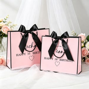 Può includere: Due sacchetti regalo rosa con bordi e manici neri, ciascuno ornato da un grande fiocco di raso nero. Le borse presentano la scritta "Happy 35 Birth" e "Happy 18th Birthday" con un simbolo a cuore.