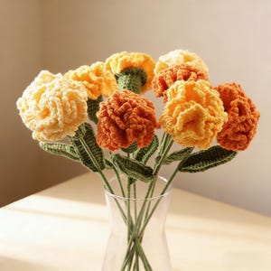 Puede incluir: Un ramo de flores de ganchillo hechas a mano en un jarrón de cristal transparente. Las flores son en tonos de amarillo, naranja y crema, con tallos y hojas verdes. Las flores tienen un aspecto texturizado y esponjoso.